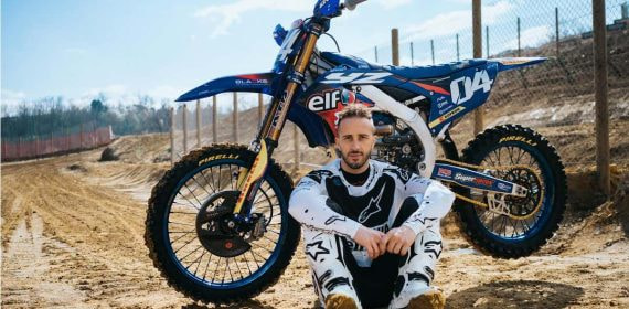 Andrea Dovizioso posa sentado junto a su moto de motocross en un circuito de tierra
