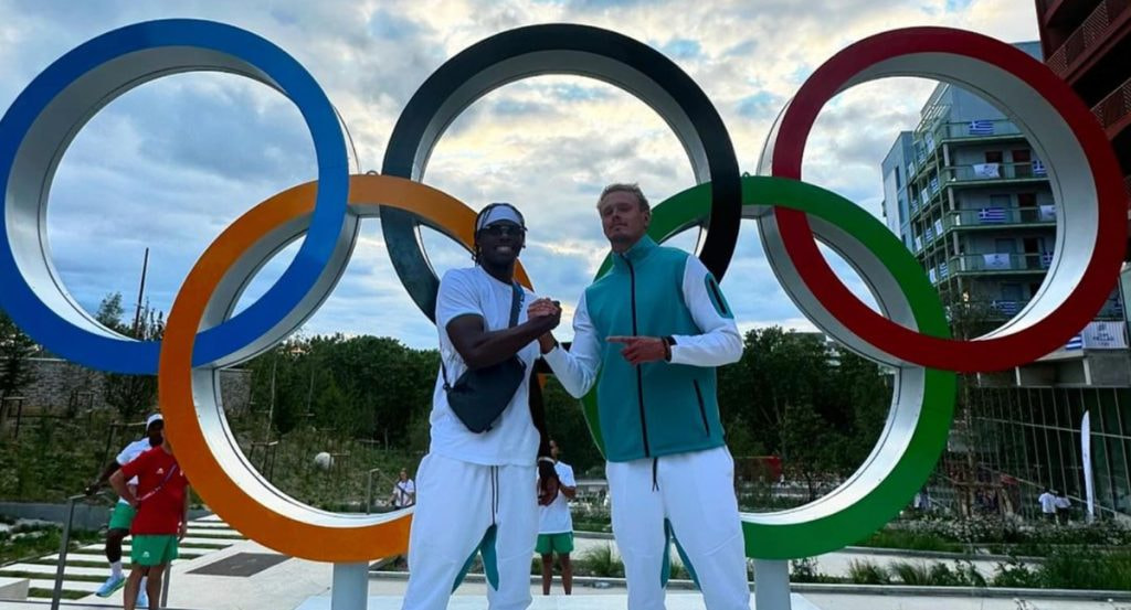 Dos deportistas se estrechan la mano ante los anillos olímpicos de la Villa de los Juegos de París 2024