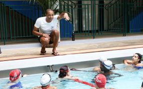 Michael Gunning, en cuclillas al borde de la piscina, da indicaciones a un grupo de jóvenes nadadores