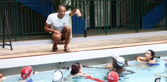 Michael Gunning, en cuclillas al borde de la piscina, da indicaciones a un grupo de jóvenes nadadores