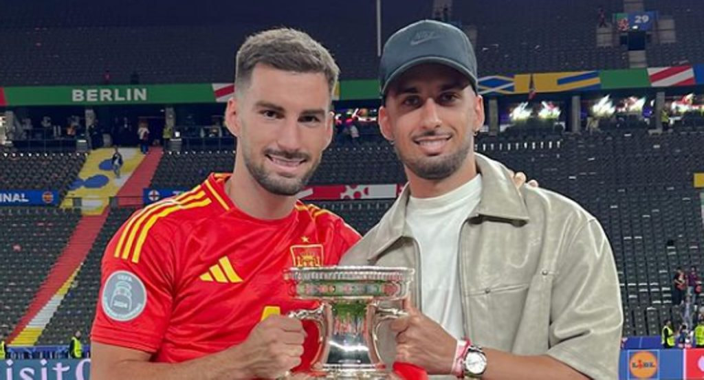 Álex Baena, con la camiseta de España, sostiene el trofeo de la Eurocopa en el estadio Olímpico de Berlín junto a un acompañante
