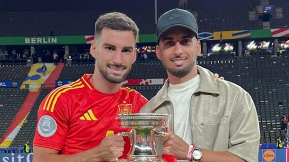 Álex Baena, con la camiseta de España, sostiene el trofeo de la Eurocopa en el estadio Olímpico de Berlín junto a un acompañante