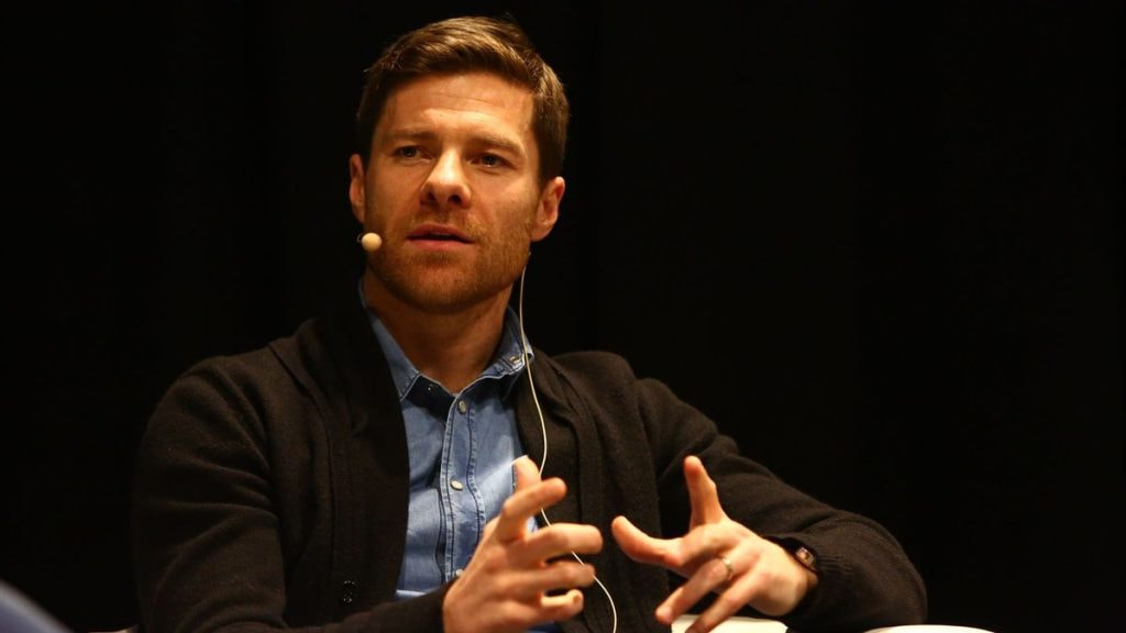 Xabi Alonso gesticula con las manos mientras habla sentado en una charla, con micrófono de diadema y camisa vaquera