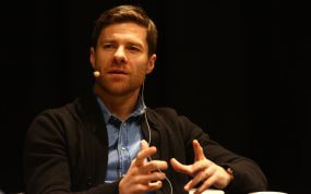 Xabi Alonso gesticula con las manos mientras habla sentado en una charla, con micrófono de diadema y camisa vaquera
