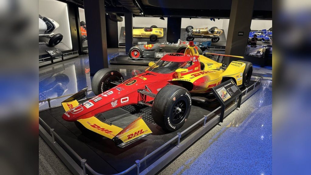 Un monoplaza de IndyCar expuesto en un museo, con coches históricos al fondo