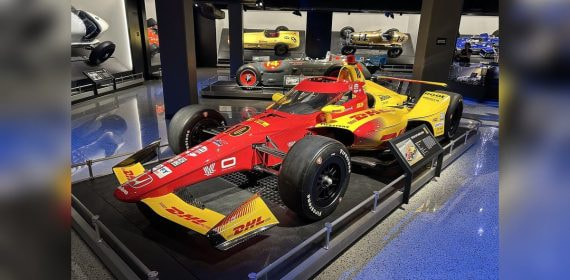 Un monoplaza de IndyCar expuesto en un museo, con coches históricos al fondo