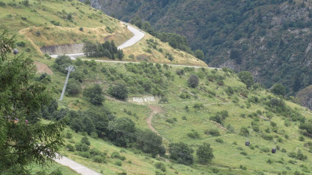 Curvas en zigzag de la subida a Alpe d'Huez entre laderas verdes con el cartel en la ladera, puerto mitico del Tour