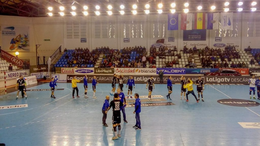 Jugadores de balonmano calientan sobre la pista antes de un partido, con la grada a medio llenar