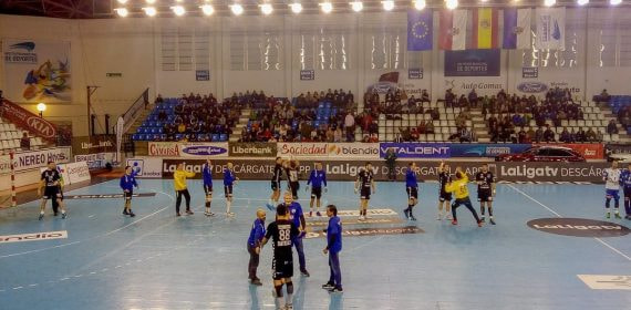 Jugadores de balonmano calientan sobre la pista antes de un partido, con la grada a medio llenar