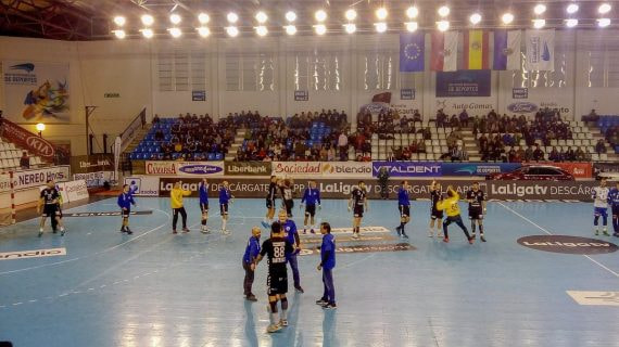 Jugadores de balonmano calientan sobre la pista antes de un partido, con la grada a medio llenar