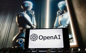 Logotipo de OpenAI (ChatGPT) en un móvil sobre un teclado y un androide al fondo mirándose en un espejo.