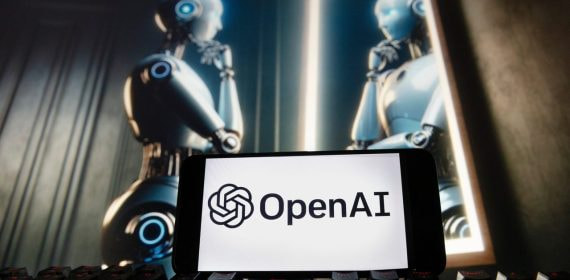 Logotipo de OpenAI (ChatGPT) en un móvil sobre un teclado y un androide al fondo mirándose en un espejo.