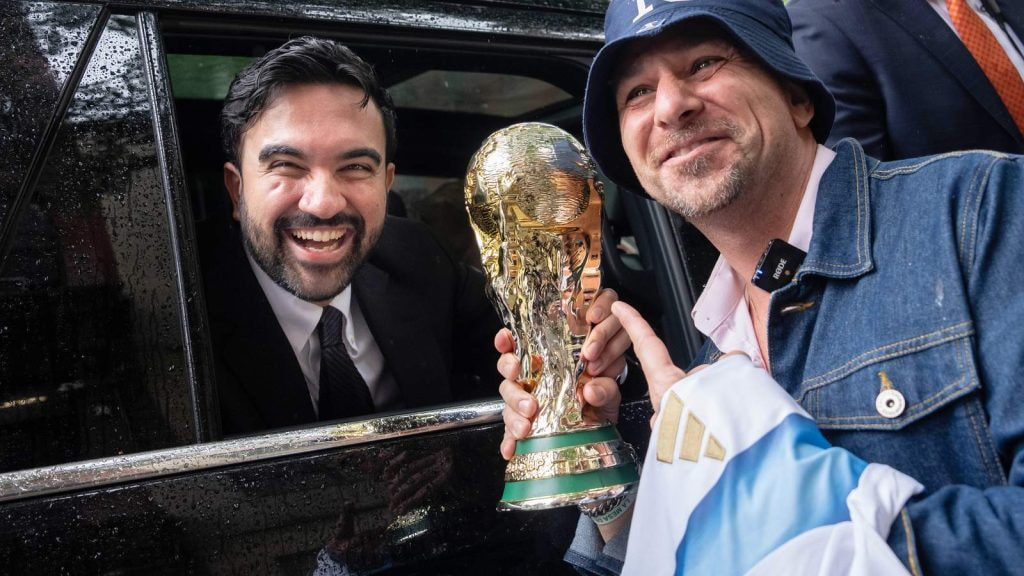Zohran Mamdani sonríe desde un coche junto a un aficionado que sostiene una réplica del trofeo del Mundial en Nueva York