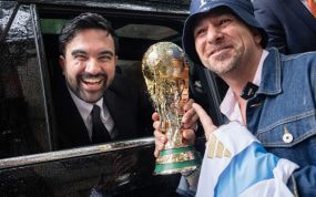 Zohran Mamdani sonríe desde un coche junto a un aficionado que sostiene una réplica del trofeo del Mundial en Nueva York