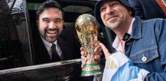 Zohran Mamdani sonríe desde un coche junto a un aficionado que sostiene una réplica del trofeo del Mundial en Nueva York