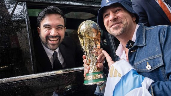 Zohran Mamdani sonríe desde un coche junto a un aficionado que sostiene una réplica del trofeo del Mundial en Nueva York