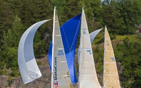 Flota de veleros de regata navegando con spinnakers desplegados junto a una costa boscosa durante una competicion de vela