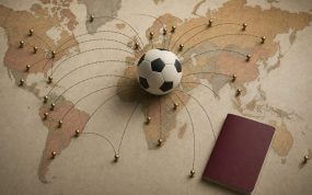 Balón de fútbol en el centro de un mapamundi con chinchetas doradas unidas por líneas hacia varios países