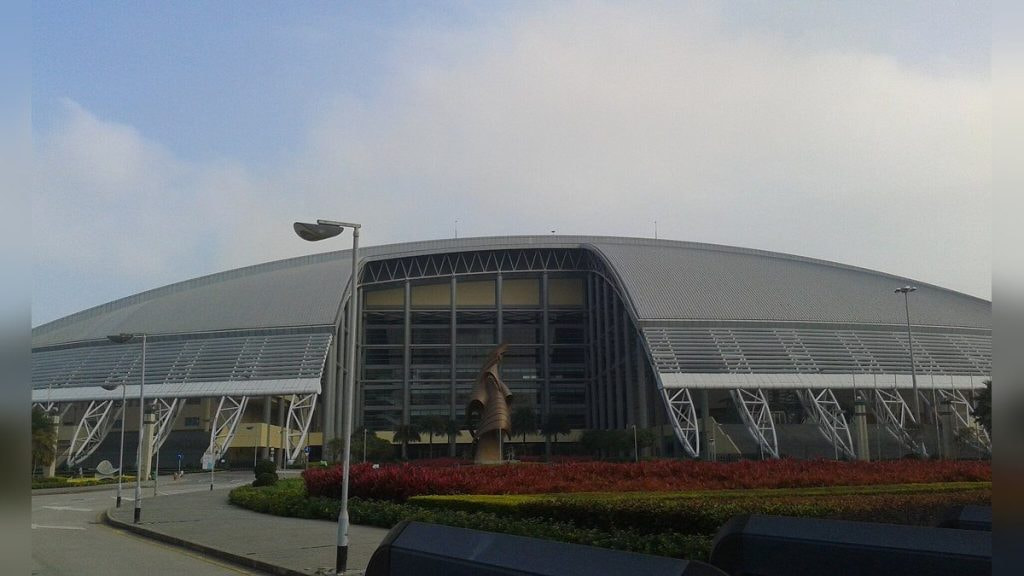 Vista exterior del pabellón cubierto de Macao, de cubierta curva gris, con los jardines y la avenida de acceso vacía