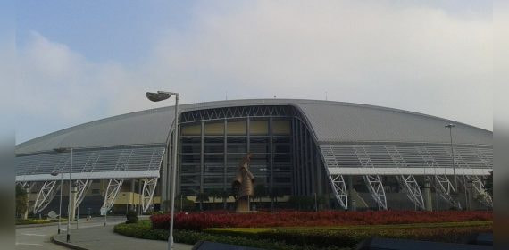 Vista exterior del pabellón cubierto de Macao, de cubierta curva gris, con los jardines y la avenida de acceso vacía