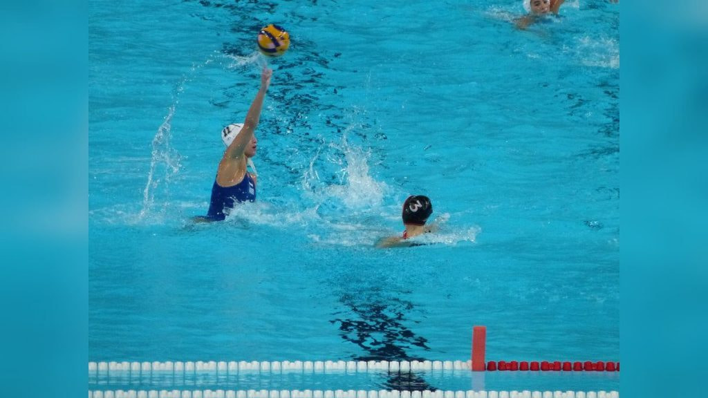 Una jugadora de waterpolo con gorro blanco arma el lanzamiento con el balón en alto ante una rival en la piscina