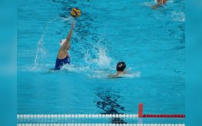 Una jugadora de waterpolo con gorro blanco arma el lanzamiento con el balón en alto ante una rival en la piscina