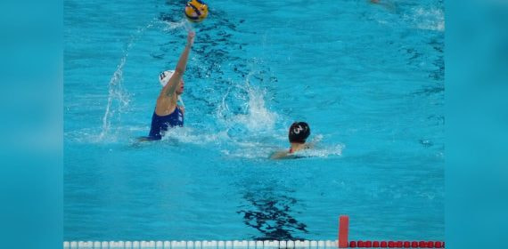 Una jugadora de waterpolo con gorro blanco arma el lanzamiento con el balón en alto ante una rival en la piscina