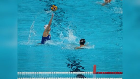 Una jugadora de waterpolo con gorro blanco arma el lanzamiento con el balón en alto ante una rival en la piscina