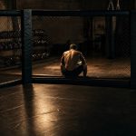 Un luchador de espaldas sentado al borde de una jaula de MMA en un gimnasio vacío de noche