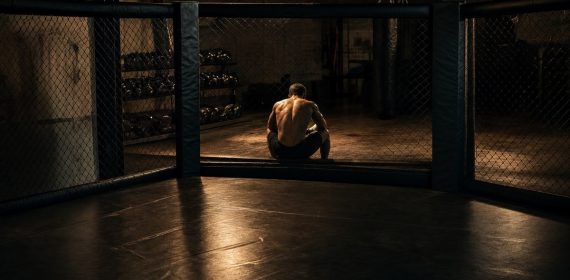 Un luchador de espaldas sentado al borde de una jaula de MMA en un gimnasio vacío de noche