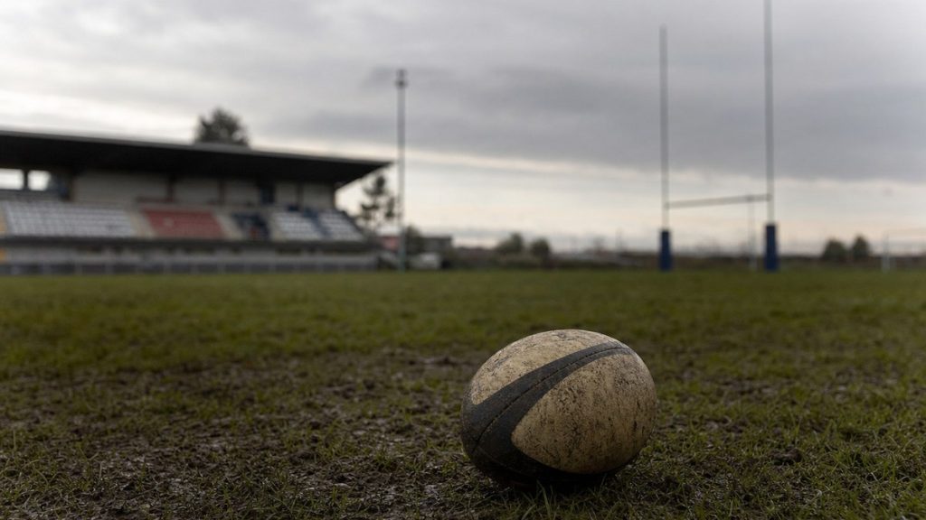 Un balón de rugby embarrado sobre el césped de un campo vacío, con las gradas y los postes al fondo.