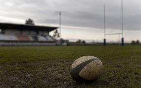 Un balón de rugby embarrado sobre el césped de un campo vacío, con las gradas y los postes al fondo.