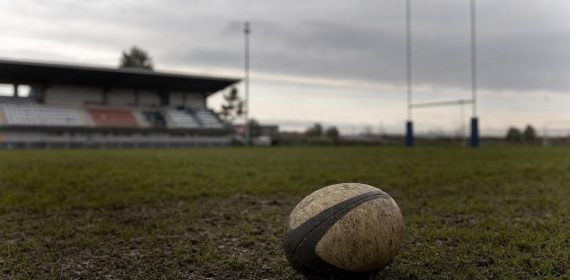 Un balón de rugby embarrado sobre el césped de un campo vacío, con las gradas y los postes al fondo.