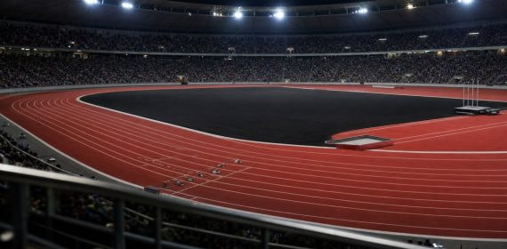Pista de atletismo iluminada de noche con el césped interior en negro y las gradas llenas de público