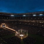 Estadio de fútbol lleno al anochecer con un ring de lucha libre iluminado en el centro del campo