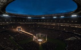Estadio de fútbol lleno al anochecer con un ring de lucha libre iluminado en el centro del campo