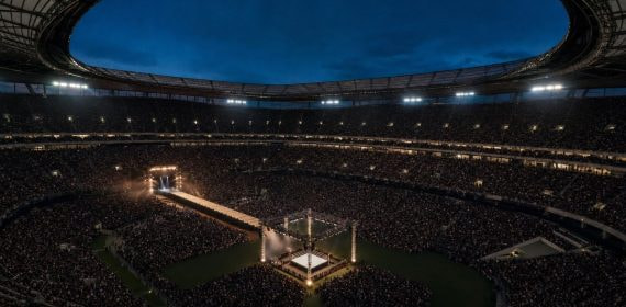 Estadio de fútbol lleno al anochecer con un ring de lucha libre iluminado en el centro del campo
