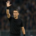 Un árbitro de fútbol levanta el brazo derecho con la mano abierta y los cinco dedos extendidos sobre el césped de un estadio con público