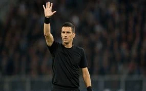 Un árbitro de fútbol levanta el brazo derecho con la mano abierta y los cinco dedos extendidos sobre el césped de un estadio con público