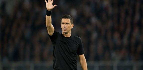 Un árbitro de fútbol levanta el brazo derecho con la mano abierta y los cinco dedos extendidos sobre el césped de un estadio con público