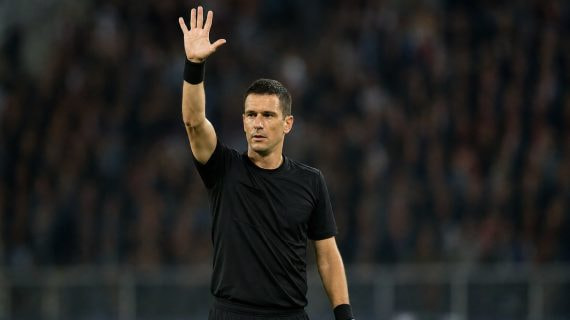 Un árbitro de fútbol levanta el brazo derecho con la mano abierta y los cinco dedos extendidos sobre el césped de un estadio con público