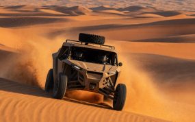 Prototipo de rally-raid sin distintivos coronando una duna y levantando una nube de arena en el desierto