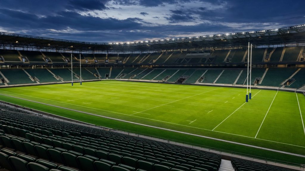 Estadio de rugby vacío e iluminado al atardecer, con la portería y el césped marcados y las gradas sin público