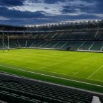 Estadio de rugby vacío e iluminado al atardecer, con la portería y el césped marcados y las gradas sin público