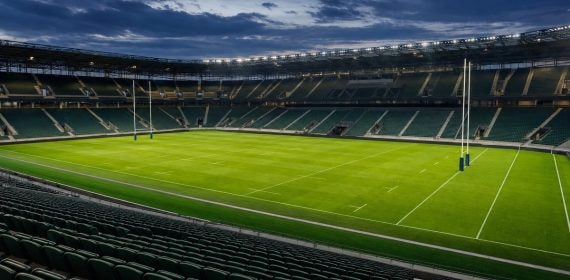 Estadio de rugby vacío e iluminado al atardecer, con la portería y el césped marcados y las gradas sin público