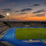 Vista general de un estadio de atletismo al atardecer, con la pista azul iluminada y las gradas llenas de público.