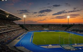 Vista general de un estadio de atletismo al atardecer, con la pista azul iluminada y las gradas llenas de público.