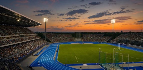 Vista general de un estadio de atletismo al atardecer, con la pista azul iluminada y las gradas llenas de público.