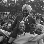 Fotografía histórica en blanco y negro de jugadores indios de hockey llevando a hombros a un directivo con turbante entre el público