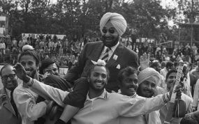 Fotografía histórica en blanco y negro de jugadores indios de hockey llevando a hombros a un directivo con turbante entre el público